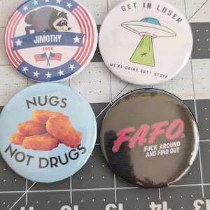 May include: Four circular pin-back buttons with various designs. One features a raccoon with the text JIMOTHY 2028, one shows a UFO with GET IN LOSER WE'RE DOING BUTT STUFF, one displays chicken nuggets with NUGS NOT DRUGS, and one says FAFO FUCK AROUND AND FIND OUT.
