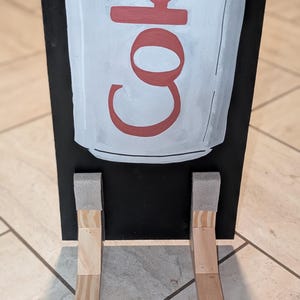 May include: A rectangular black wooden sign featuring a hand-painted light gray soda can graphic with the red text Diet Coke. The sign is supported by two light-colored wooden L-shaped brackets attached to the bottom front, each with a gray foam pad.