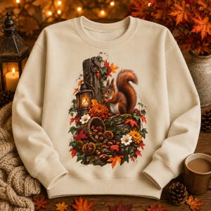 May include: A beige crewneck sweatshirt featuring a central graphic illustration of a brown squirrel perched on a mossy log next to a lit lantern, surrounded by autumn leaves, orange and white flowers, and a basket filled with acorns and chestnuts.