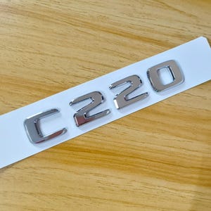Könnte beinhalten: Ein verchromtes Auto-Emblem mit dem alphanumerischen Text C220. Die einzelnen Buchstaben und Zahlen sind auf einem weißen Klebestreifen montiert und zeichnen sich durch eine reflektierende Metalloberfläche mit abgeschrägten Kanten aus.