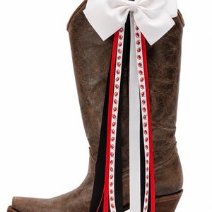 May include: A brown distressed leather cowboy boot adorned with a large white hair bow at the top. Several long ribbons hang from the bow, featuring solid black, solid red, and white ribbons patterned with small red football graphics.
