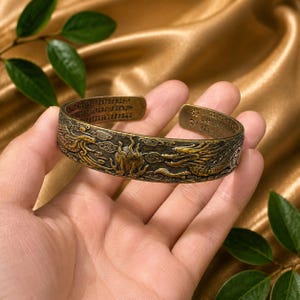 May include: A brass-toned metal cuff bracelet featuring an embossed design of a dragon and a flaming pearl amidst clouds. The interior and edges of the open-ended band are engraved with intricate Chinese characters throughout the entire surface.