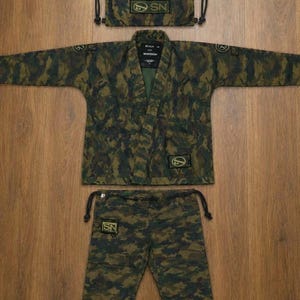 May include: A camouflage print jiu-jitsu gi set consisting of a long-sleeve jacket, matching pants with a drawstring waist, and a drawstring storage bag. Each piece features black rectangular patches with the text SN and a circular logo.