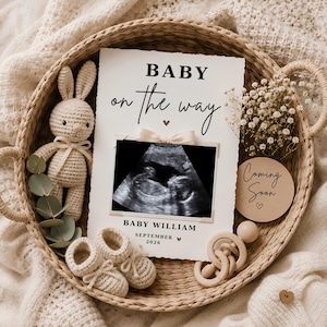Puede incluir: Una cesta tejida que contiene una tarjeta de anuncio blanca con el texto BABY on the way, BABY WILLIAM y SEPTEMBER 2026, una imagen de ecografía, un conejo de ganchillo, un par de patucos de ganchillo, un sonajero de madera y flores secas.