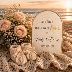 May include: An arched sign with the text "And Then There Were Four Baby Williams OCTOBER 2026" next to a bouquet of pale pink roses and baby's breath, and a pair of beige knitted baby booties resting on a cream-colored crocheted fabric.