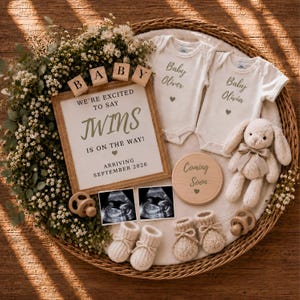 Puede incluir: Una cesta de mimbre que contiene dos bodies blancos para bebé con las etiquetas Baby Oliver y Baby Olivia, un cartel que dice We're Excited To Say Twins Is On The Way Arriving September 2026, dos ecografías, un conejo de peluche, dos chupetes y patucos.