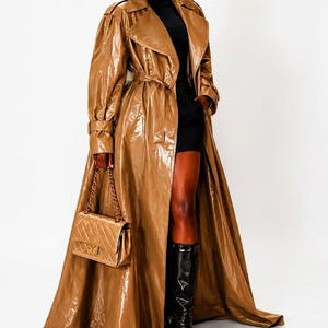 Könnte beinhalten: Ein langer, glänzender, hellbrauner Trenchcoat mit breitem Kragen, langen Ärmeln und einem passenden Taillengürtel. Der Mantel hat eine lackartige Oberfläche. Eine rechteckige, gesteppte hellbraune Handtasche mit goldener Kette.