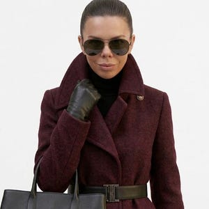 May include: A deep burgundy wool-blend coat featuring a wide collar, a black turtleneck sweater underneath, a black leather belt with a rectangular buckle, black leather gloves, and a black structured handbag, paired with dark aviator sunglasses.