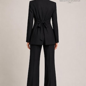 May include: A black suit consisting of a blazer and wide-leg trousers. The blazer features a notched collar, long sleeves with buttons at the cuffs, and a fabric tie at the waist in the back. The text STUDIOELARALLC EST. 2024 is visible.