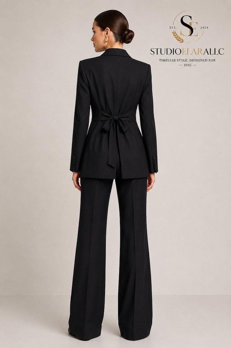 May include: A black suit consisting of a blazer and wide-leg trousers. The blazer features a notched collar, long sleeves with buttons at the cuffs, and a fabric tie at the waist in the back. The text STUDIOELARALLC EST. 2024 is visible.