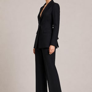 May include: A black tailored suit consisting of a single-button blazer with notched lapels and matching wide-leg trousers. The blazer features flap pockets and long sleeves. The logo STUDIOELARALLC EST. 2024 TIMELESS STYLE, DESIGNED FOR YOU is visible.