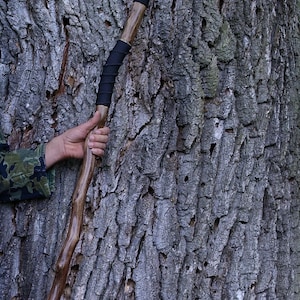 May include: A wooden walking stick with a natural, gnarled brown finish, featuring a clear spherical topper and black cord wrapping around the upper section. The base of the stick is fitted with a black rubber tip for stability and grip.