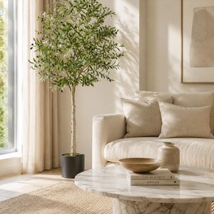May include: A tall artificial olive tree with green leaves and small black olives in a dark cylindrical pot. The tree features a slender, textured trunk and a full, rounded canopy, standing next to a sofa and a round white marble coffee table.