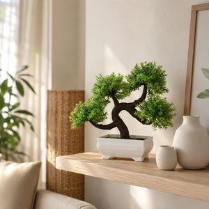 May include: An artificial bonsai tree with a dark brown, textured, curved trunk and vibrant green foliage. The tree is potted in a rectangular, white ceramic container with four small feet, positioned on a light-colored wooden shelf.