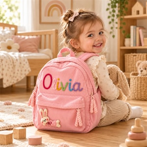 Puede incluir: Una mochila de pana rosa claro con el nombre Olivia bordado en letras coloridas en el bolsillo delantero. La bolsa incluye una pequeña aplicación de un conejo blanco y rosa, tiradores de cremallera trenzados a juego y correas ajustables.