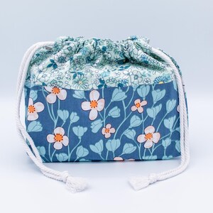 May include: A fabric drawstring pouch featuring a two-tone floral design. The top section has a light blue background with small white flowers, while the bottom section is a darker blue with larger white and light blue flowers with orange centers.