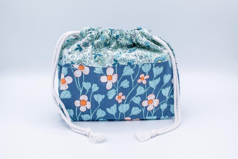 May include: A fabric drawstring pouch featuring a two-tone floral design. The top section has a light blue background with small white flowers, while the bottom section is a darker blue with larger white and light blue flowers with orange centers.