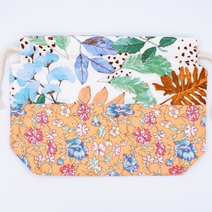 May include: A rectangular fabric drawstring pouch featuring a two-part design. The top section displays a white background with blue, green, and brown botanical leaf illustrations. The bottom section has an orange background with small pink and blue floral patterns. Two cream-coloured rope cords are attached to the sides.