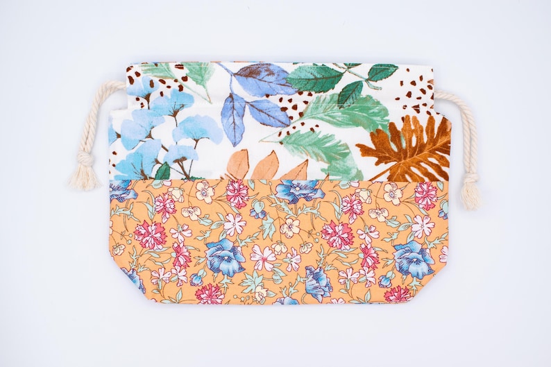May include: A rectangular fabric drawstring pouch featuring a two-part design. The top section displays a white background with blue, green, and brown botanical leaf illustrations. The bottom section has an orange background with small pink and blue floral patterns. Two cream-coloured rope cords are attached to the sides.