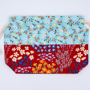May include: A rectangular fabric drawstring pouch featuring a two-tone floral design. The top section is light blue with small white and yellow flowers, while the bottom section is deep red with larger yellow, white, and blue floral patterns.