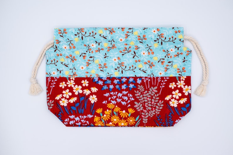May include: A rectangular fabric drawstring pouch featuring a two-tone floral design. The top section is light blue with small white and yellow flowers, while the bottom section is deep red with larger yellow, white, and blue floral patterns.