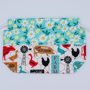May include: A drawstring pouch featuring a top section with a white daisy pattern on a teal background and a bottom section with a farm-themed print on a white background, including illustrations of chickens, pigs, geese, and windmills.