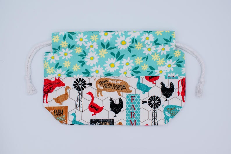 May include: A drawstring pouch featuring a top section with a white daisy pattern on a teal background and a bottom section with a farm-themed print on a white background, including illustrations of chickens, pigs, geese, and windmills.