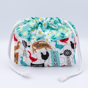 May include: A fabric drawstring pouch featuring a top section with a white daisy pattern on a teal background and a bottom section with a farm-themed print including chickens, geese, pigs, and windmills on a white background with wire fencing.