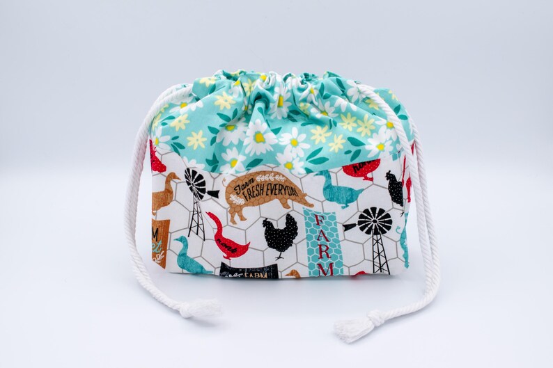 May include: A fabric drawstring pouch featuring a top section with a white daisy pattern on a teal background and a bottom section with a farm-themed print including chickens, geese, pigs, and windmills on a white background with wire fencing.