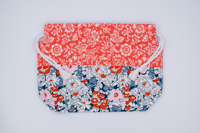 May include: A rectangular drawstring pouch featuring a two-tone floral pattern. The top section displays a vibrant coral-orange background with white botanical motifs, while the bottom section showcases a dense floral print in navy, coral, and pink.