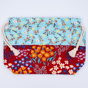 May include: A rectangular fabric drawstring pouch featuring two distinct floral patterns. The top section is light blue with small orange berries and white flowers, while the bottom section is deep red with yellow and white wildflower prints.