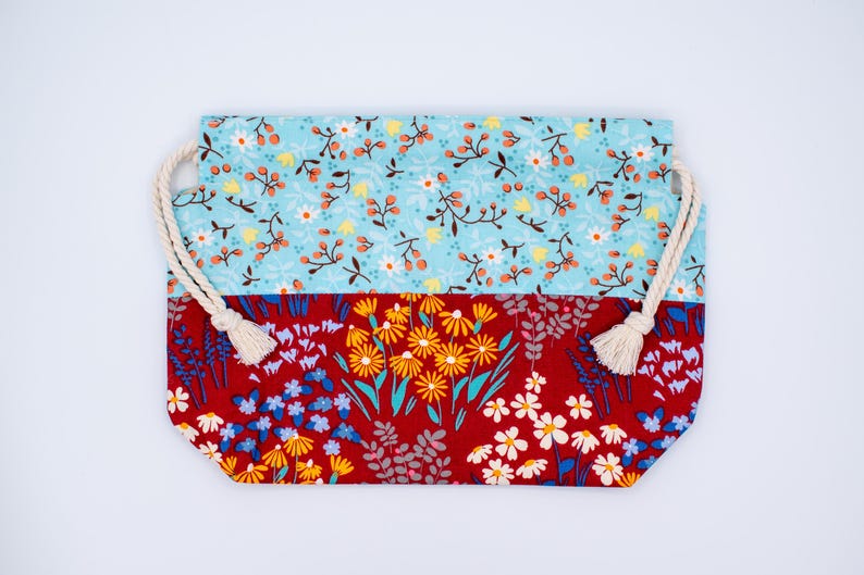 May include: A rectangular fabric drawstring pouch featuring two distinct floral patterns. The top section is light blue with small orange berries and white flowers, while the bottom section is deep red with yellow and white wildflower prints.