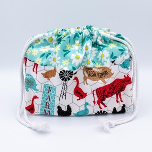 May include: A fabric drawstring pouch featuring a top section with a white and yellow daisy pattern on a teal background. The lower section displays a farm-themed print with chickens, a cow, a pig, a windmill, and the text FARM FRESH EVERYDAY.