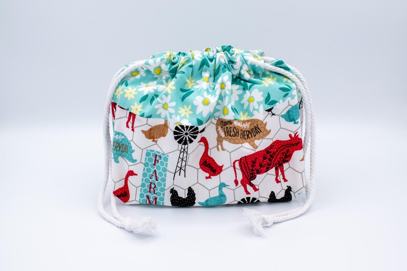 May include: A fabric drawstring pouch featuring a top section with a white and yellow daisy pattern on a teal background. The lower section displays a farm-themed print with chickens, a cow, a pig, a windmill, and the text FARM FRESH EVERYDAY.