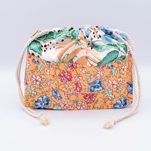 May include: A fabric drawstring pouch featuring a two-tone design. The lower section displays an orange background with a dense floral pattern of pink and blue flowers. The upper section is white with green leaves and brown stems, finished with white rope.