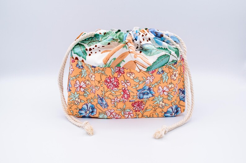 May include: A fabric drawstring pouch featuring a two-tone design. The lower section displays an orange background with a dense floral pattern of pink and blue flowers. The upper section is white with green leaves and brown stems, finished with white rope.
