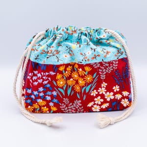 May include: A drawstring pouch featuring a two-tone design. The top section is light blue with a small floral pattern, while the bottom section is red with a vibrant print of yellow, white, and blue wildflowers. It has thick white rope cords.