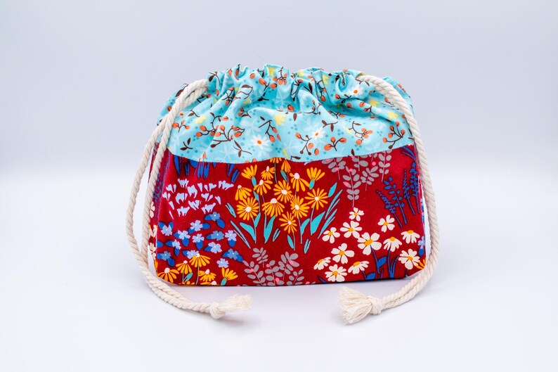 May include: A drawstring pouch featuring a two-tone design. The top section is light blue with a small floral pattern, while the bottom section is red with a vibrant print of yellow, white, and blue wildflowers. It has thick white rope cords.