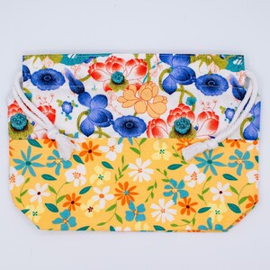 Puede incluir: Una bolsa rectangular de tela con cord&oacute;n y dise&ntilde;o bicolor. La parte superior muestra un fondo blanco con motivos de flores de loto azules, rojas y naranjas. La parte inferior es amarilla con un estampado de peque&ntilde;as flores blancas, naranjas y azules.