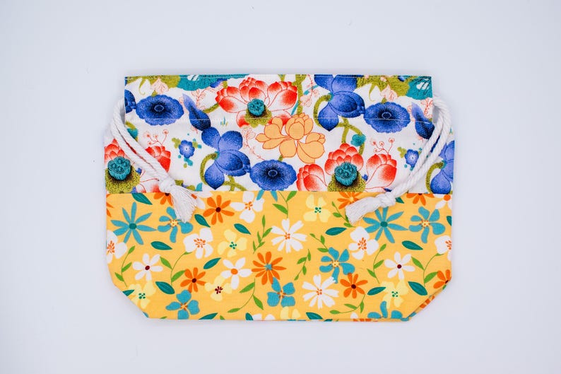 Puede incluir: Una bolsa rectangular de tela con cord&oacute;n y dise&ntilde;o bicolor. La parte superior muestra un fondo blanco con motivos de flores de loto azules, rojas y naranjas. La parte inferior es amarilla con un estampado de peque&ntilde;as flores blancas, naranjas y azules.