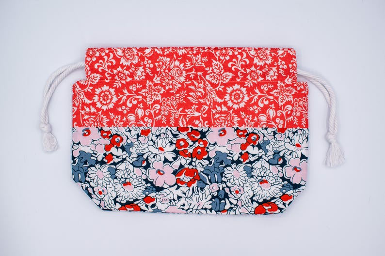 May include: A rectangular drawstring pouch featuring two distinct floral patterns. The top section displays a vibrant red fabric with a white botanical print, while the bottom section showcases a dense floral design in red, pink, and dark blue.