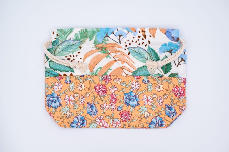 May include: A rectangular fabric pouch featuring a two-tone design. The top section displays a white background with green leaves and orange fronds. The bottom section features an orange background with a dense floral pattern of blue and pink flowers.
