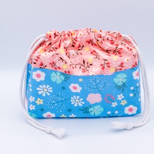 May include: A fabric drawstring pouch featuring a two-tone design. The upper section is a soft peach color with a delicate floral pattern, while the lower section is a bright blue with a larger, colorful floral print. It has white rope cords.