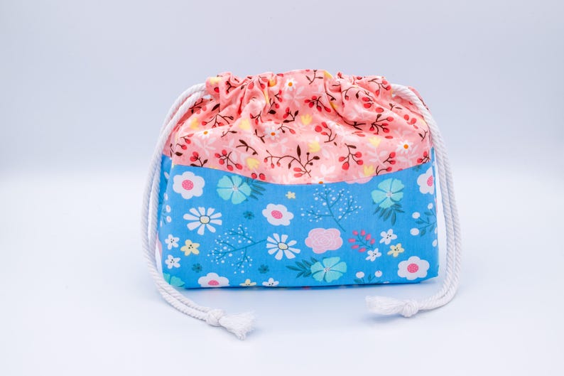 May include: A fabric drawstring pouch featuring a two-tone design. The upper section is a soft peach color with a delicate floral pattern, while the lower section is a bright blue with a larger, colorful floral print. It has white rope cords.