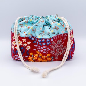 May include: A fabric drawstring pouch featuring a two-tone design. The top section is light blue with a small floral pattern, while the bottom section is deep red with a larger print of white, yellow, and blue flowers and green foliage.