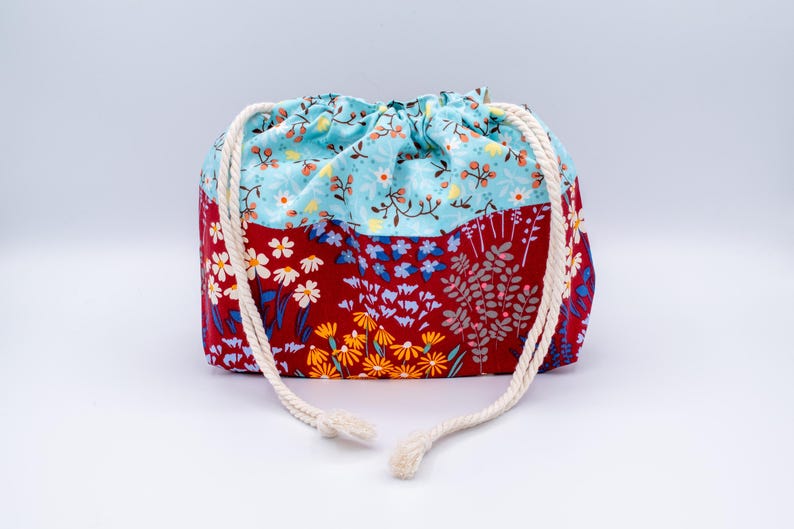 May include: A fabric drawstring pouch featuring a two-tone design. The top section is light blue with a small floral pattern, while the bottom section is deep red with a larger print of white, yellow, and blue flowers and green foliage.