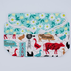 May include: A fabric drawstring pouch featuring a top section with a white daisy pattern on a teal background and a bottom section with a farm-themed print on a white background, including cows, pigs, geese, windmills, and the text Farm Fresh Everyday.
