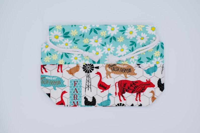 May include: A fabric drawstring pouch featuring a top section with a white daisy pattern on a teal background and a bottom section with a farm-themed print on a white background, including cows, pigs, geese, windmills, and the text Farm Fresh Everyday.