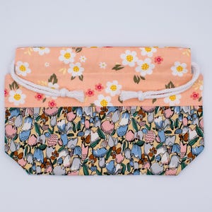 Pode incluir: Uma bolsa retangular com cord&atilde;o e design floral de dois tons. A parte superior &eacute; de tecido p&ecirc;ssego claro com flores brancas e amarelas, enquanto a parte inferior exibe um padr&atilde;o denso de pequenas tulipas azuis, rosas e amarelas.