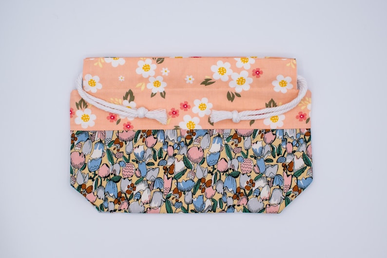Pode incluir: Uma bolsa retangular com cord&atilde;o e design floral de dois tons. A parte superior &eacute; de tecido p&ecirc;ssego claro com flores brancas e amarelas, enquanto a parte inferior exibe um padr&atilde;o denso de pequenas tulipas azuis, rosas e amarelas.